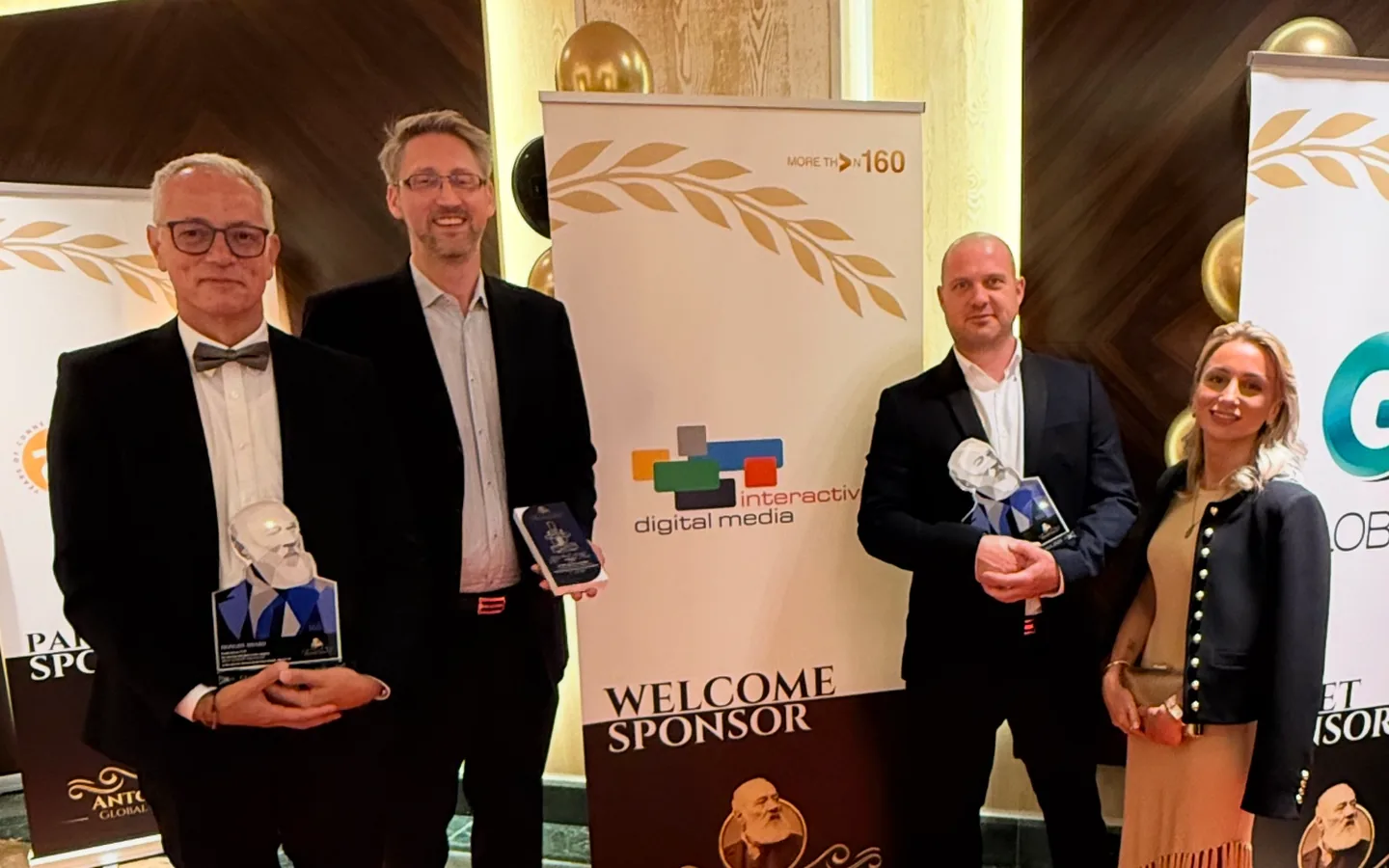 Four people holding awards on a red carpet beside a sponsor banner