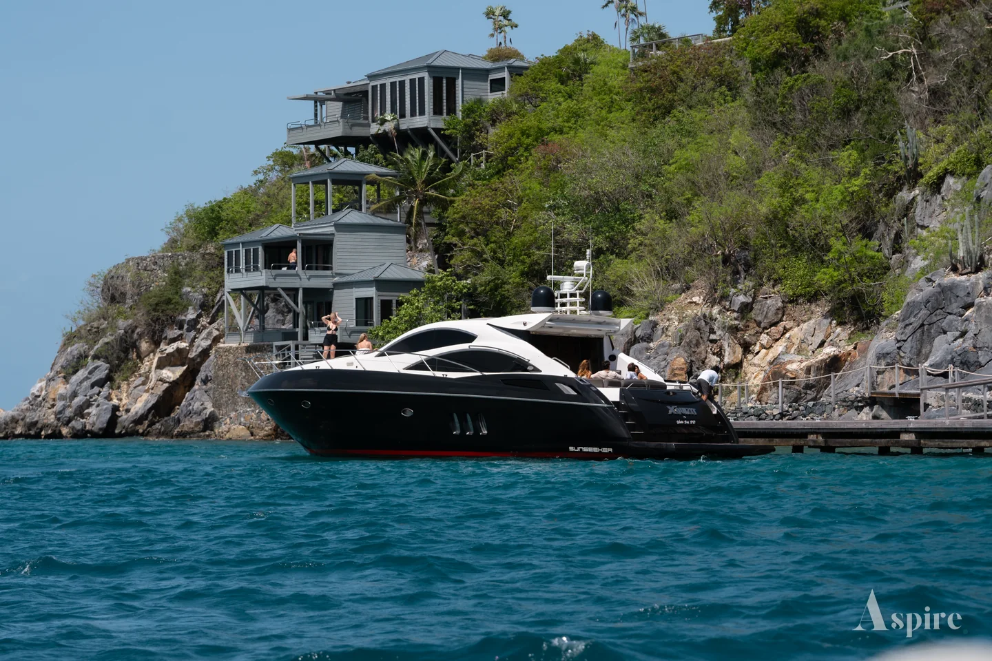 A motor yacht at anchor below a clifftop villa on a wooded headland
