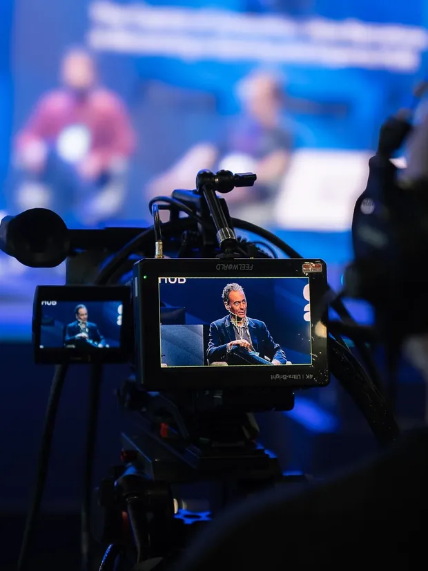 A camera on a tripod filming a speaker on stage